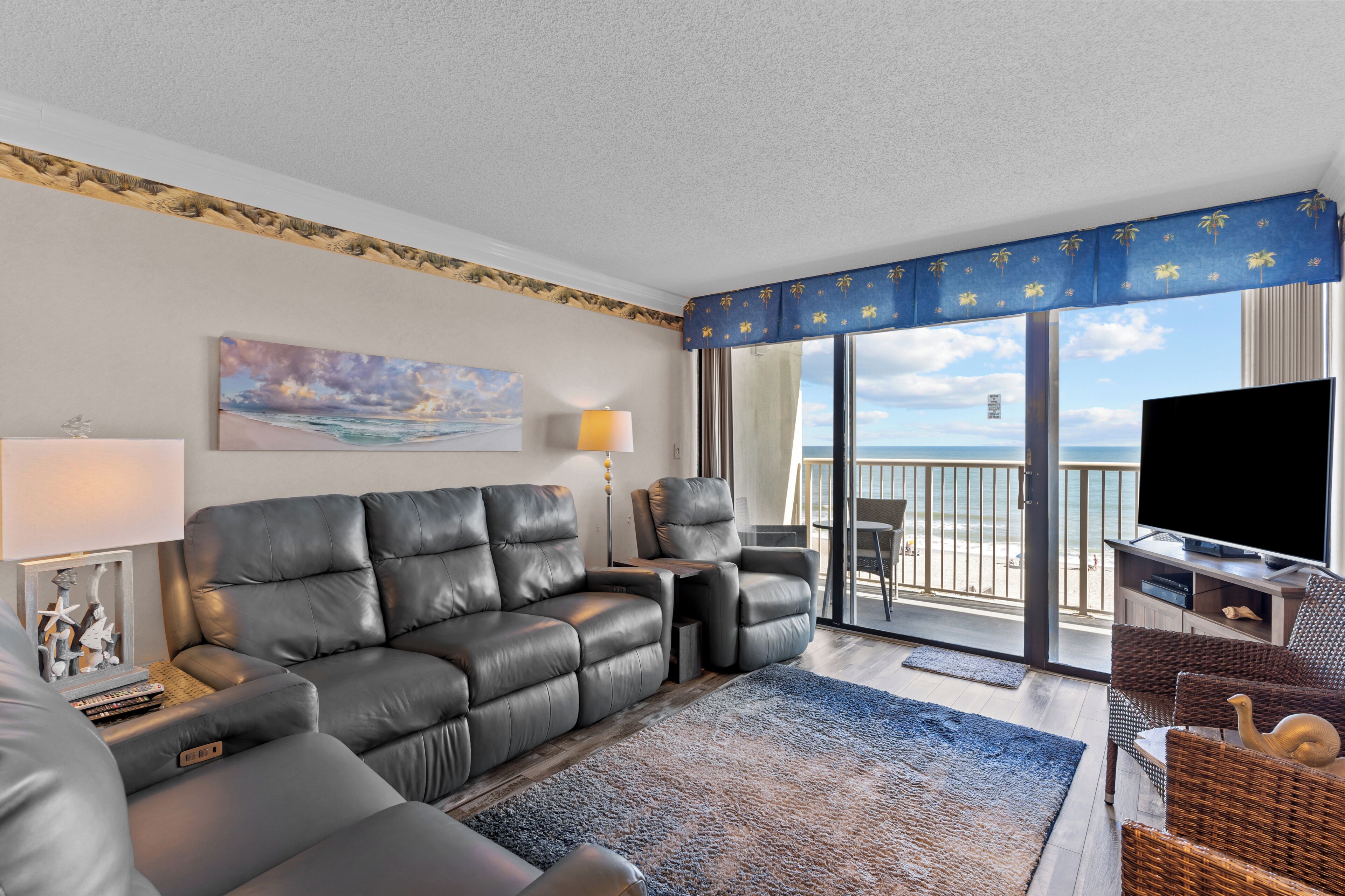 A modern living room with a leather sofa, a flat-screen TV, and a balcony with a view of the beach.
