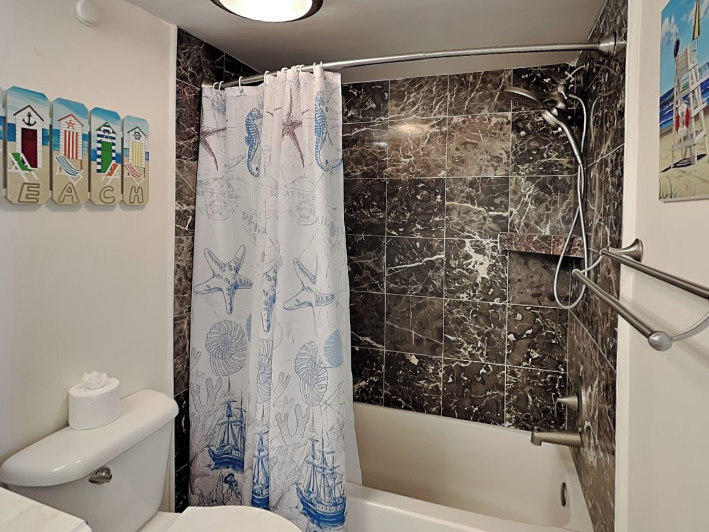 A bathroom with a shower curtain featuring nautical designs, a toilet, and a bathtub with a marble backsplash.