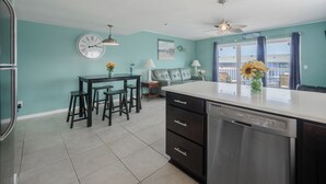 A kitchen with a large island, a dining area with stools, a sofa, and a view of the beach through a sliding door. - Sunskipper by Carolina Retreats (Carolina Beach)