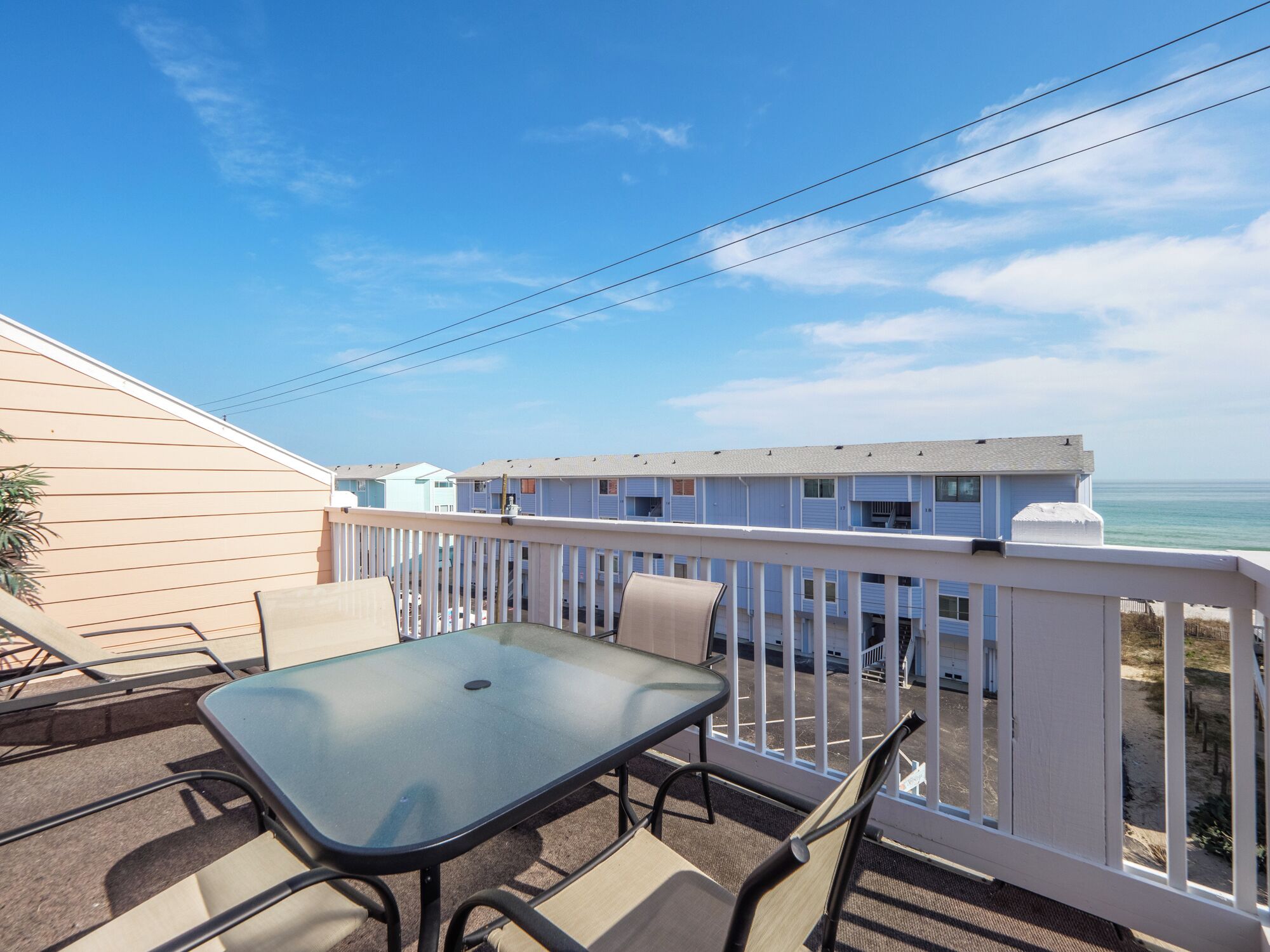A balcony with a glass table and chairs, overlooking a beach and a row of white buildings.