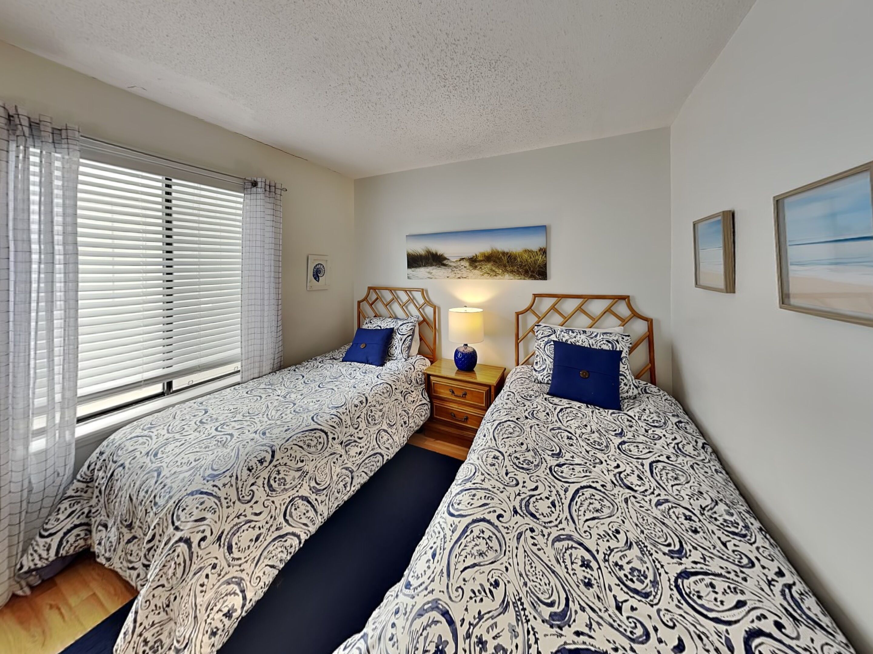 Une chambre avec deux lits, une table de chevet, une lampe et un tableau sur le thème de la plage accroché au mur.