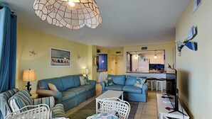 Una sala de estar con un sofá azul, una mesa de centro blanca y una silla de mimbre. Se observa una cocina al fondo con una encimera azul y armarios blancos.