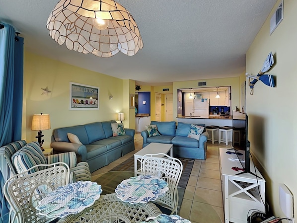 A living room with a blue sofa, a white coffee table, and a wicker chair. There is a kitchen in the background with a blue countertop and white cabinets. - St. Regis by Carolina Retreats (Sneads Ferry)