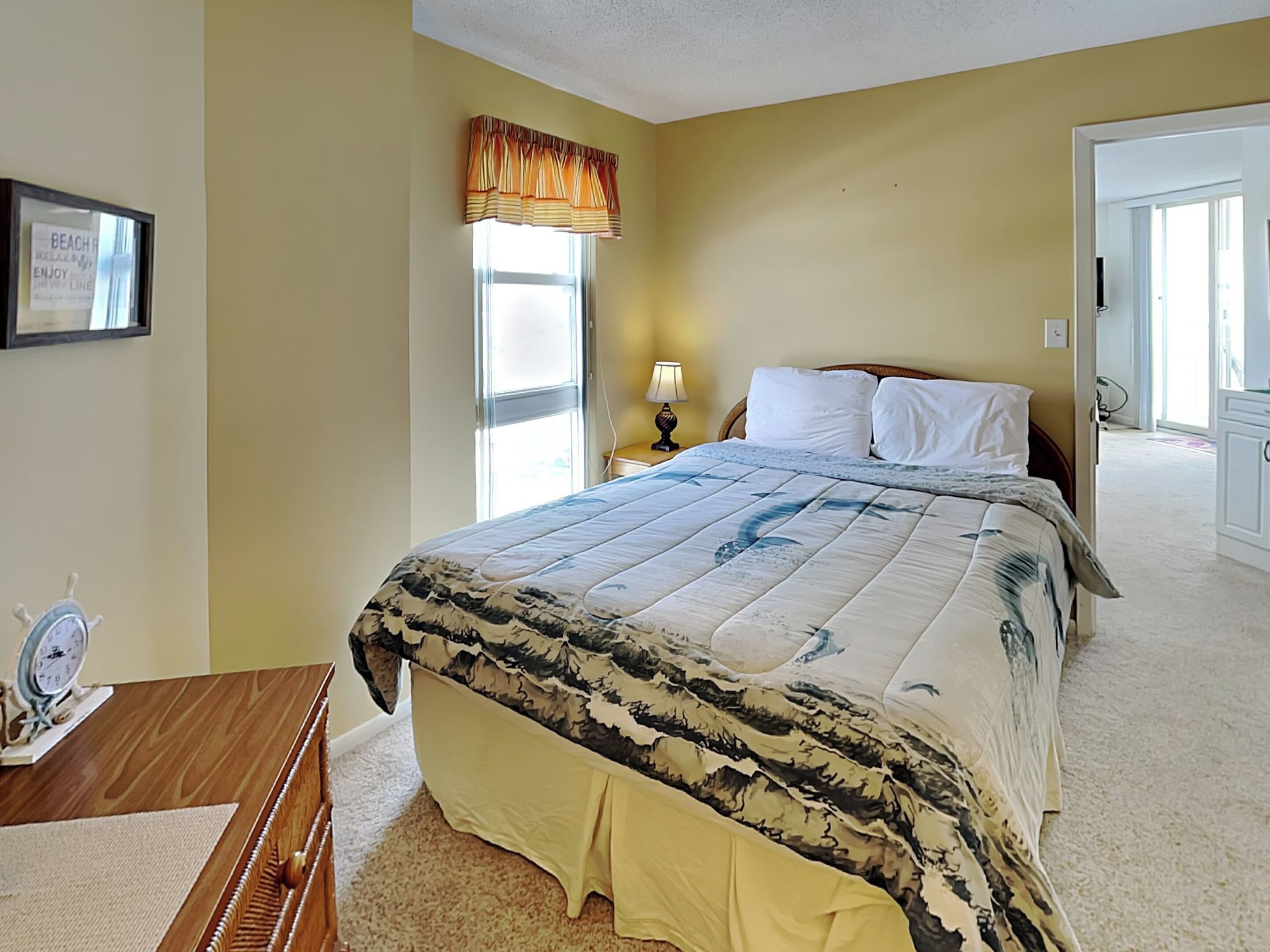 A bedroom with a bed, a nightstand, a lamp, a framed picture, and a window with curtains.