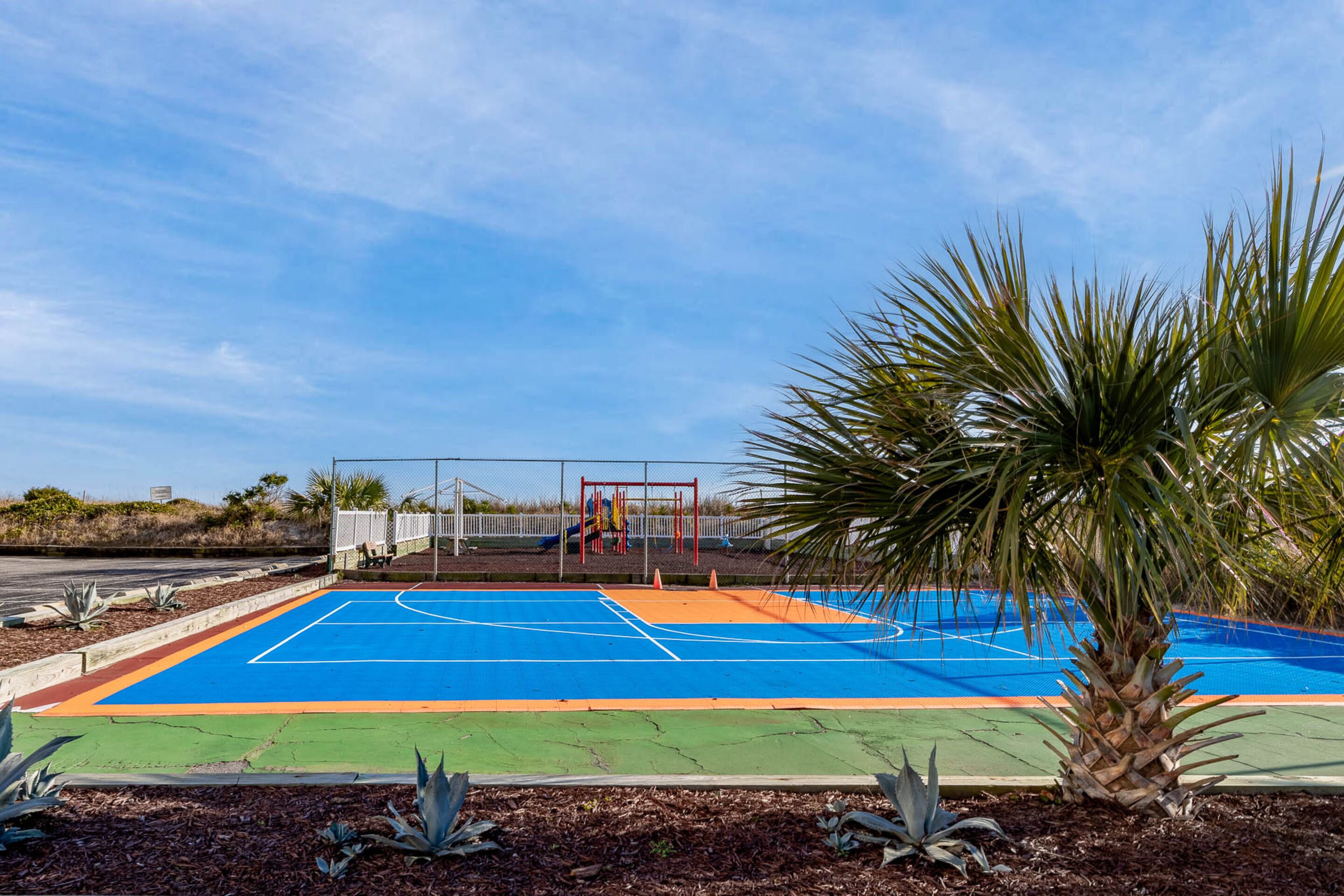 Una cancha de baloncesto al aire libre con valla, palmeras y un parque infantil al fondo.