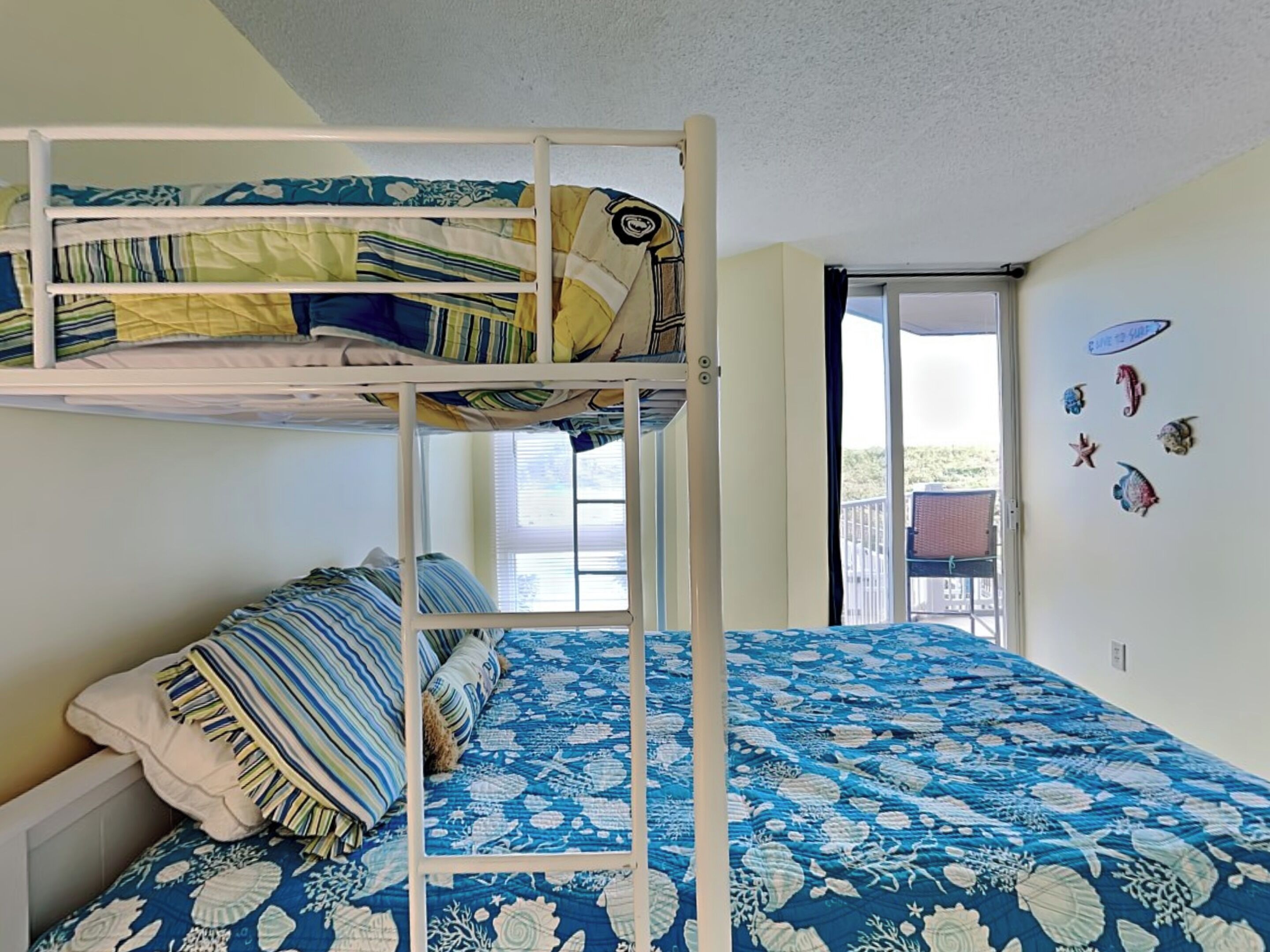 A bunk bed room with a blue patterned bedspread, a window with blinds, and a door leading to an outdoor balcony.