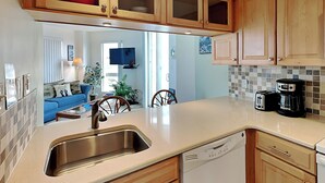 A kitchen with wooden cabinets, a stainless steel sink, and a dishwasher. There is a dining area with a blue sofa and a television in the background. - St. Regis by Carolina Retreats (Sneads Ferry)