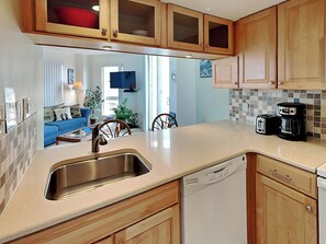 A kitchen with wooden cabinets, a stainless steel sink, and a dishwasher. There is a dining area with a blue sofa and a television in the background. - St. Regis by Carolina Retreats (Sneads Ferry)