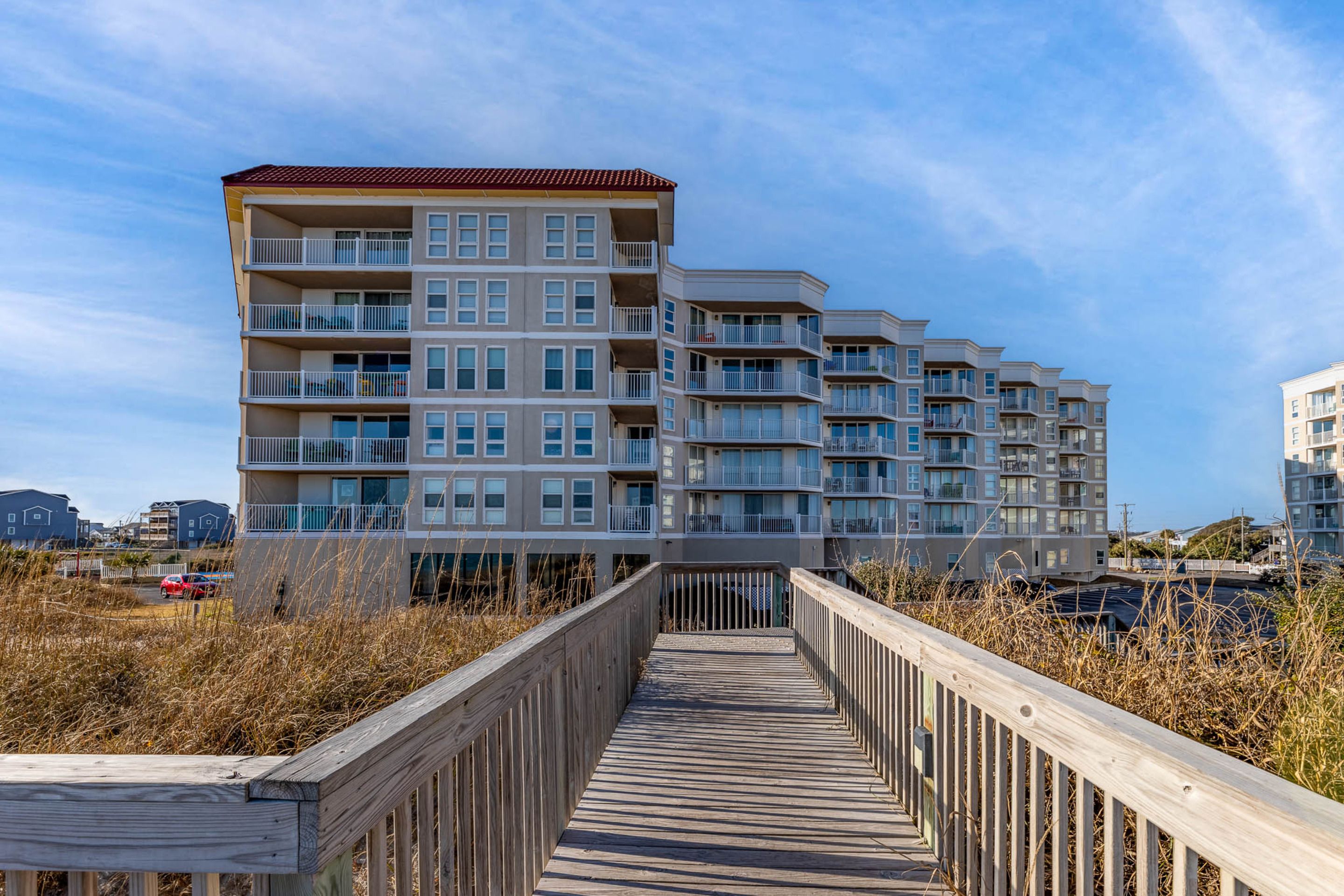 A multi-story residential building with balconies, a wooden boardwalk leading to it, and a clear sky.