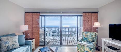Una sala de estar con sofá, sillón y mesa de centro, un mueble para la TV, y un balcón con vista al mar.