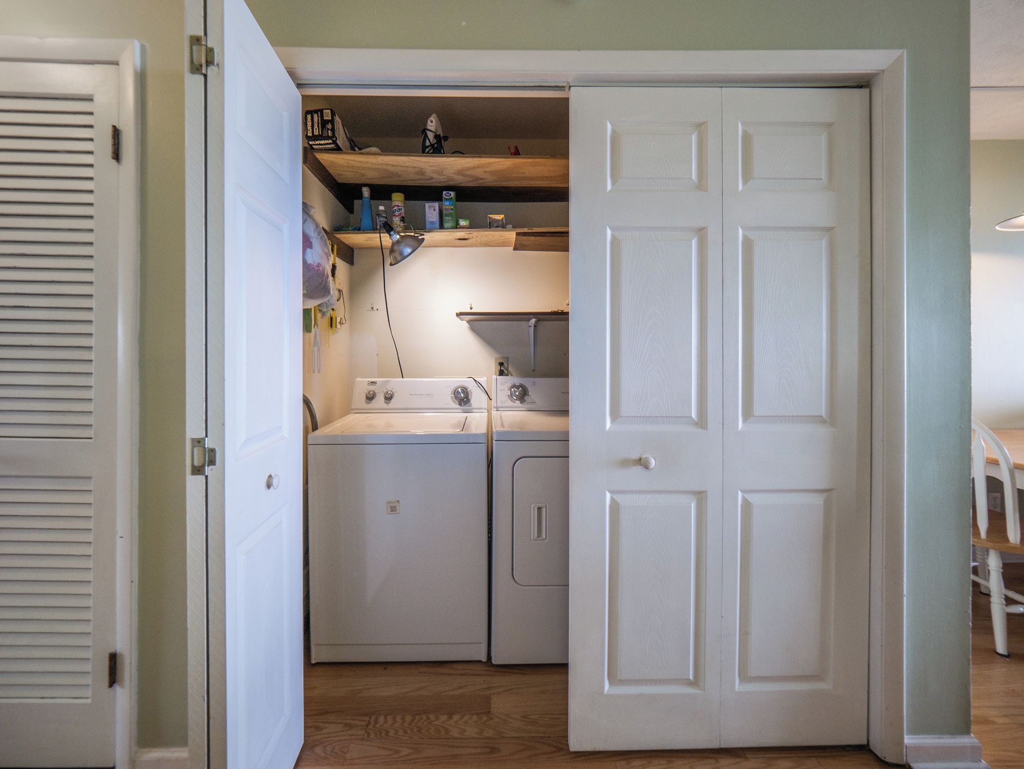 Un cuarto de lavado con dos lavadoras y secadoras apiladas, un estante con varios objetos y una puerta que da a otra habitación.