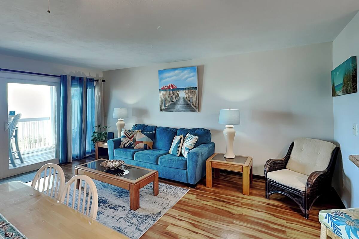 A living room with a blue sofa, a wooden coffee table, a side table with a lamp, a wicker chair, and a painting on the wall.