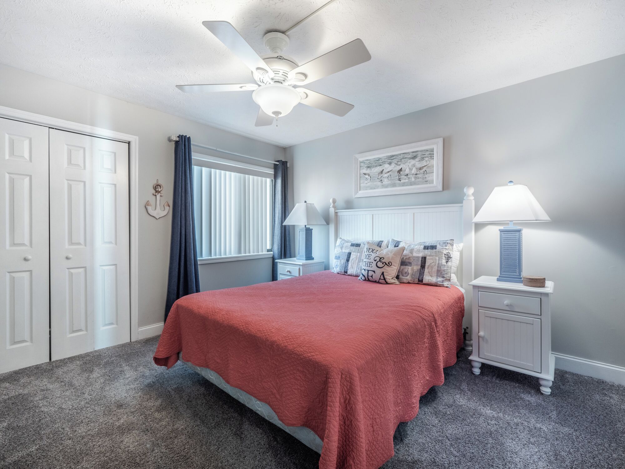 A bedroom with a bed, a ceiling fan, a nightstand with a lamp, and a window with curtains.