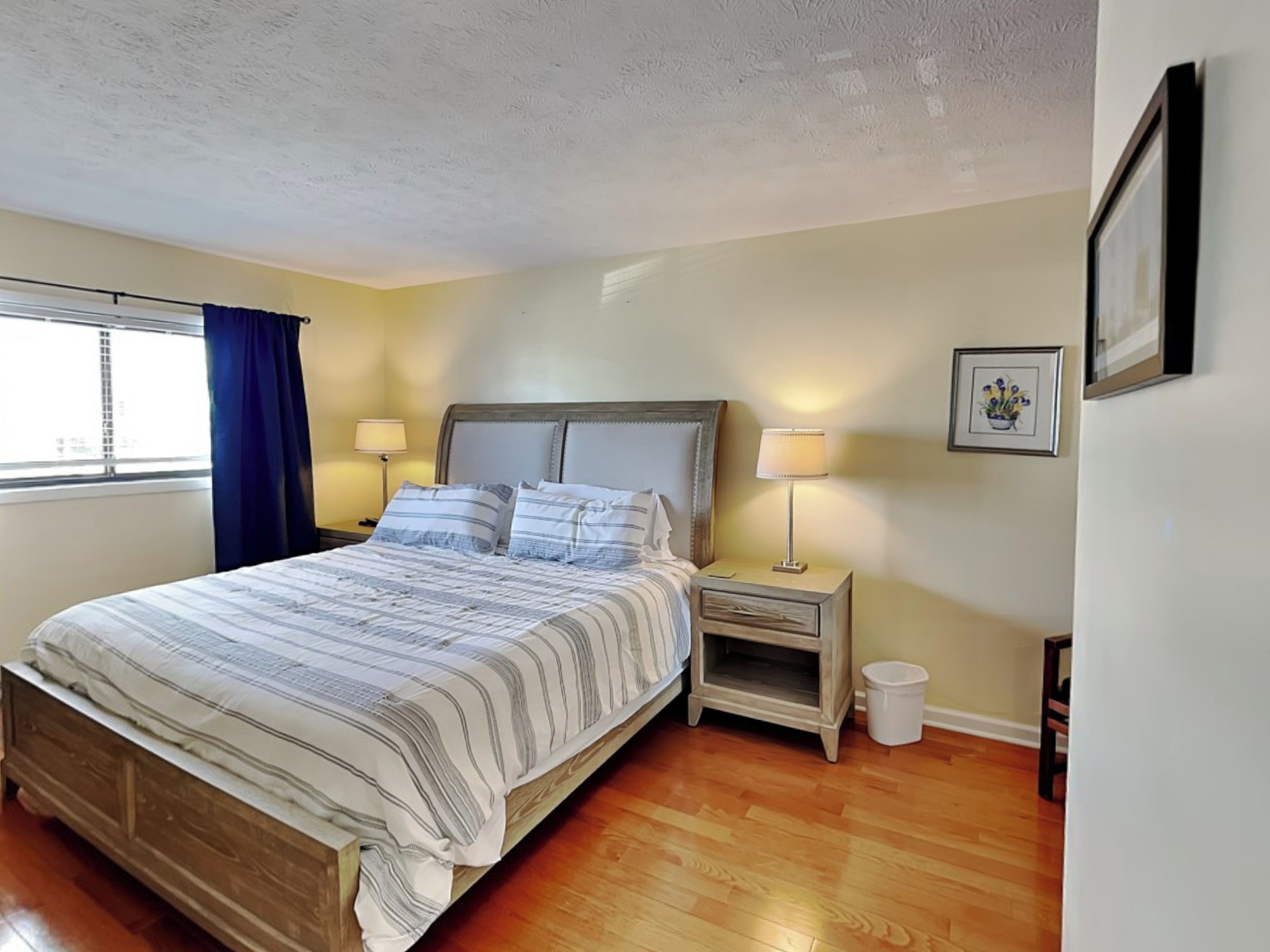 Un dormitorio con cama de madera, dos mesitas de noche con lámparas, una ventana con cortinas y un cuadro en la pared.