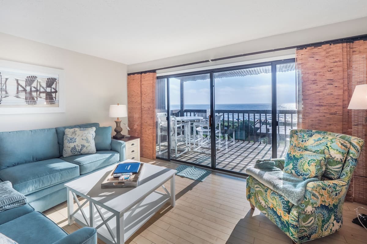 A spacious balcony with a view of the ocean, a blue sofa, a patterned armchair, and a white coffee table.