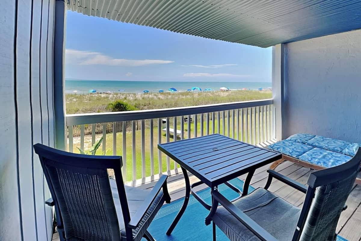 A balcony with a view of the beach, a table, and two chairs.