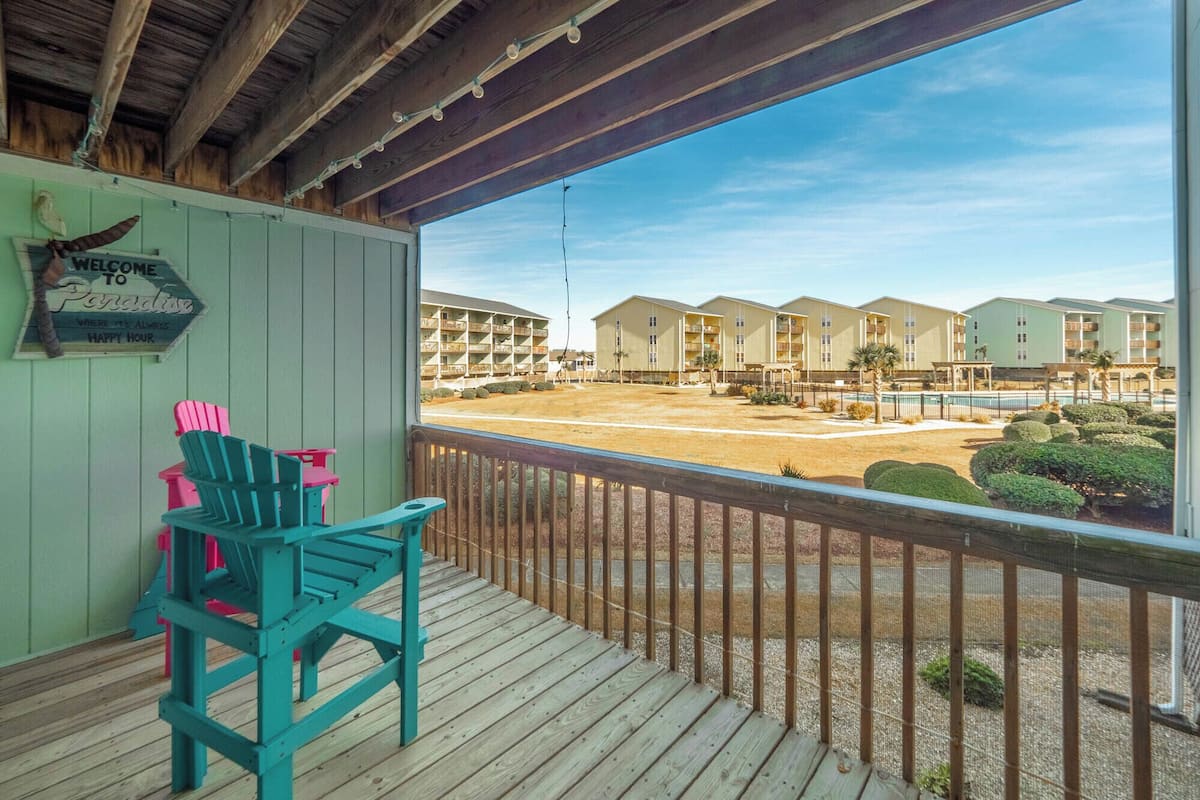 A wooden deck with turquoise chairs, a welcome sign, and a view of a resort with multiple buildings and a pool.