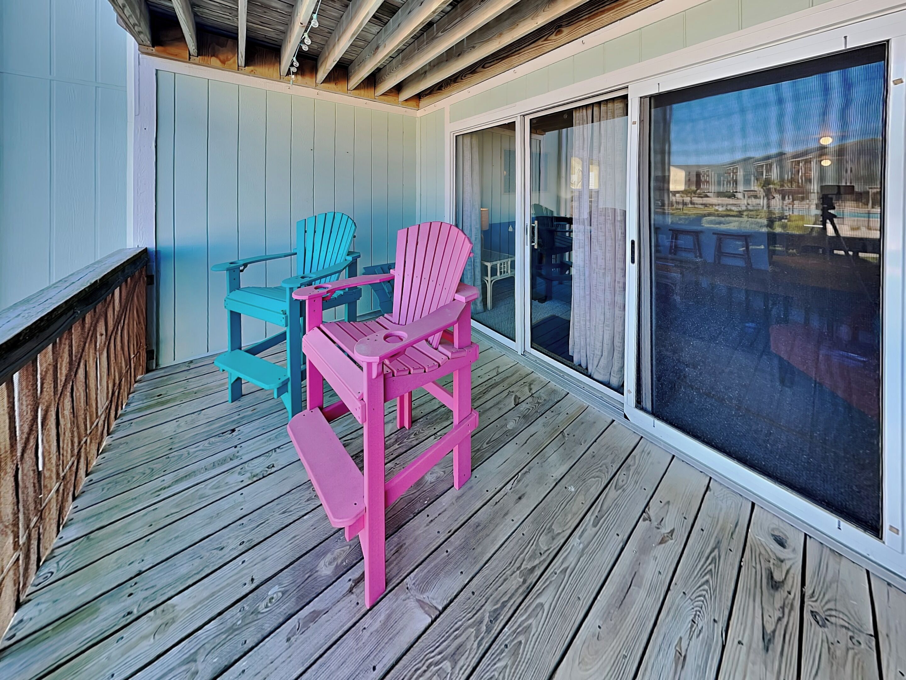 Una terraza de madera con dos sillas Adirondack, una rosa y otra azul, y un ventanal corredero que da a un patio al aire libre.