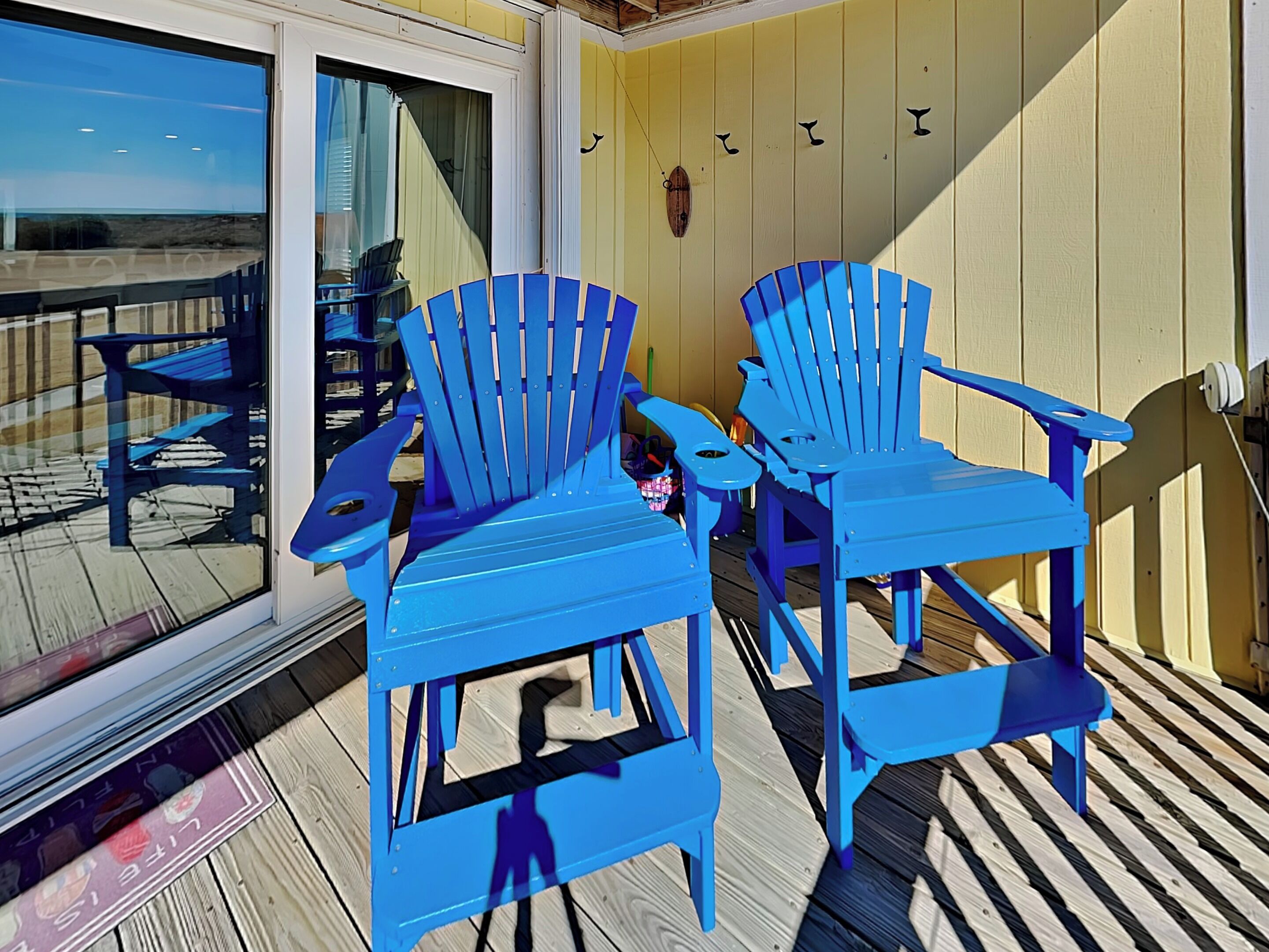 Zwei blaue Adirondack-Stühle auf einer Holzterrasse mit einer Glastür, die zu einem Balkon führt.