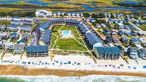 Luftaufnahme eines Küstenresorts mit mehreren Gebäuden, einem zentralen Pool und einem Strandbereich mit Sonnenschirmen.