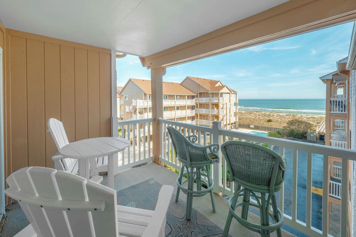 A balcony with a view of the beach and ocean, white chairs, and a small table.
