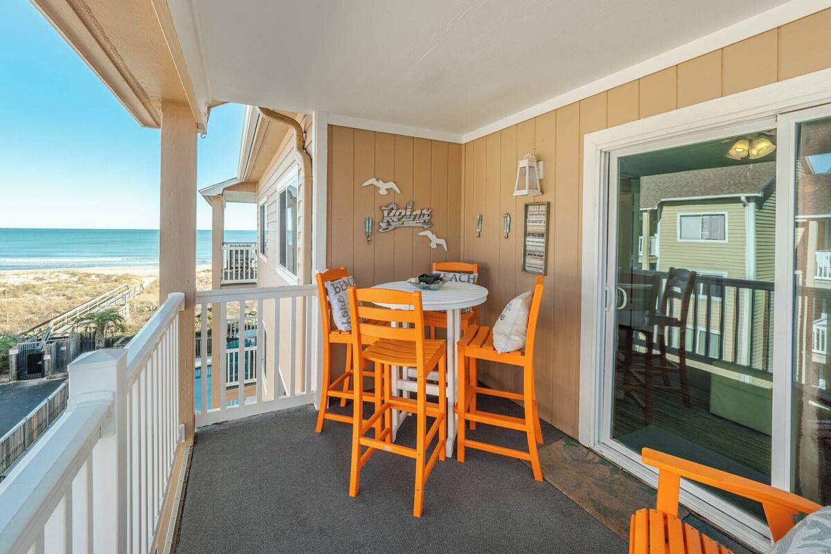 A balcony with a table and chairs, a view of the beach, and a wooden house in the background.