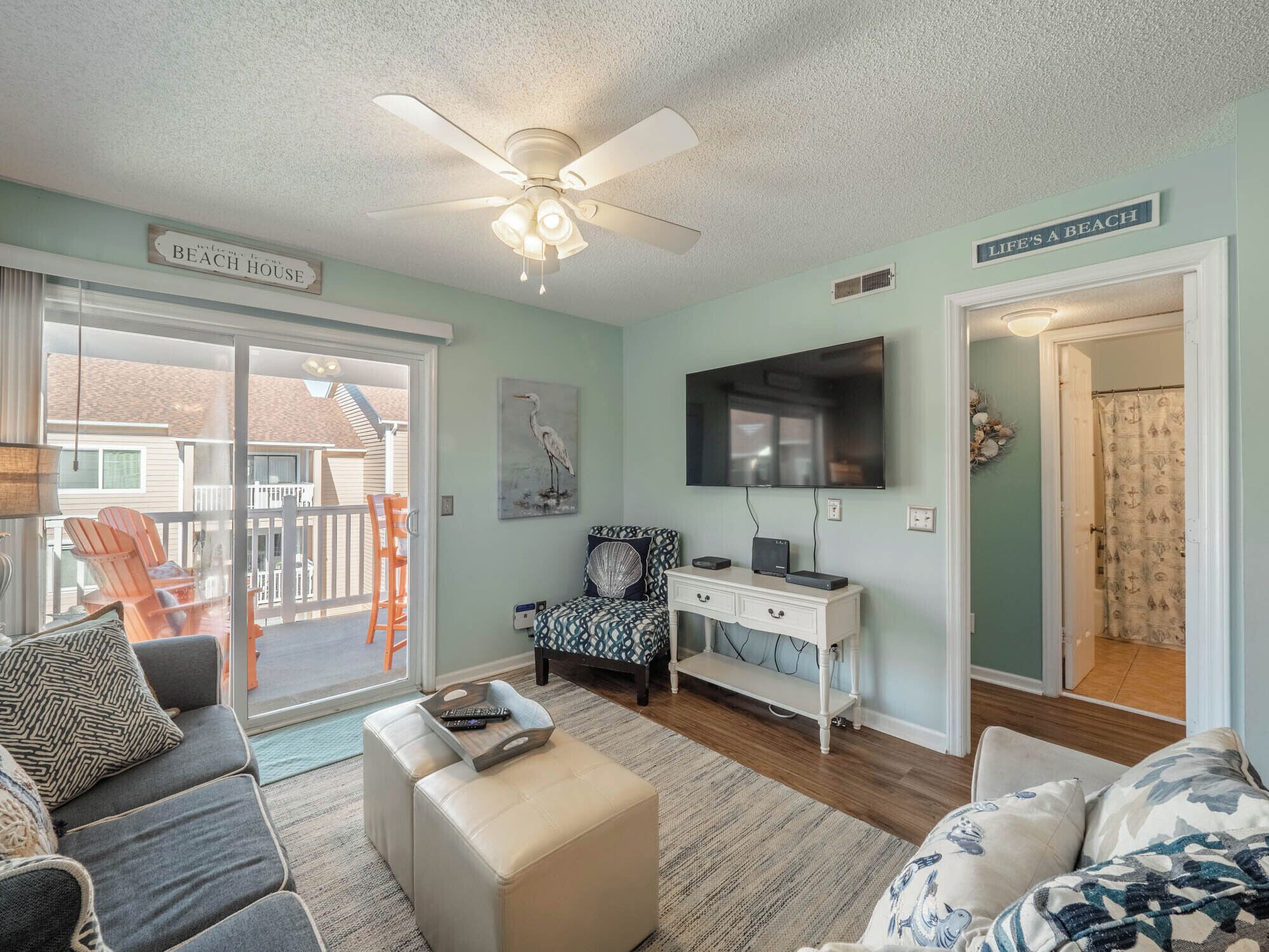 A living room with a ceiling fan, a flat-screen TV, a sofa, a coffee table, and a chair. There is a sign on the wall that reads "Welcome to our Beach House".