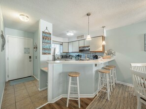 A modern kitchen with a central island, bar stools, and a view of the adjacent living area. - Sand Pebbles by Carolina Retreats (Carolina Beach)