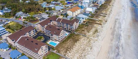 An aerial view of a coastal residential area with beachfront properties, including multi-story buildings with balconies and a beach access walkway.