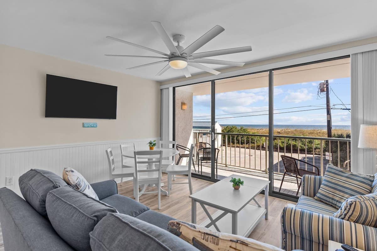 Una sala de estar moderna con ventilador de techo, televisor de pantalla plana y balcón con vista a la playa.