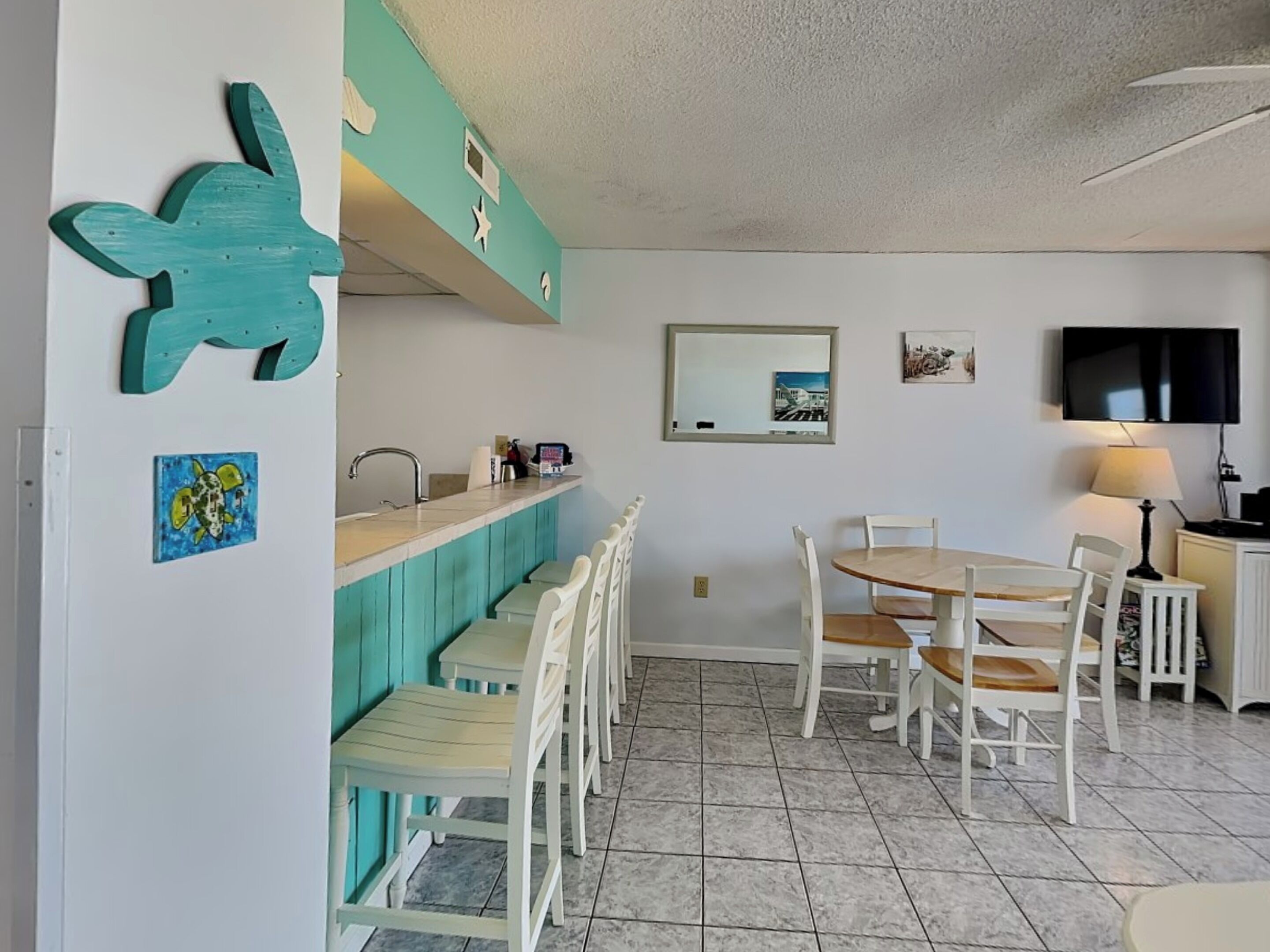 A compact kitchen with a bar area, white chairs, a dining table, and a TV mounted on the wall.