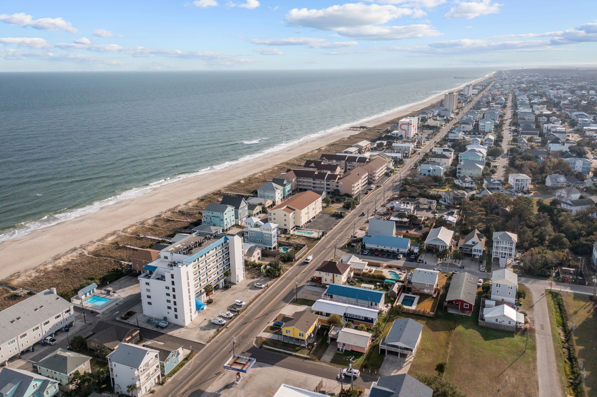 An aerial view of a coastal town with a beach, residential buildings, and a road running parallel to the shore.