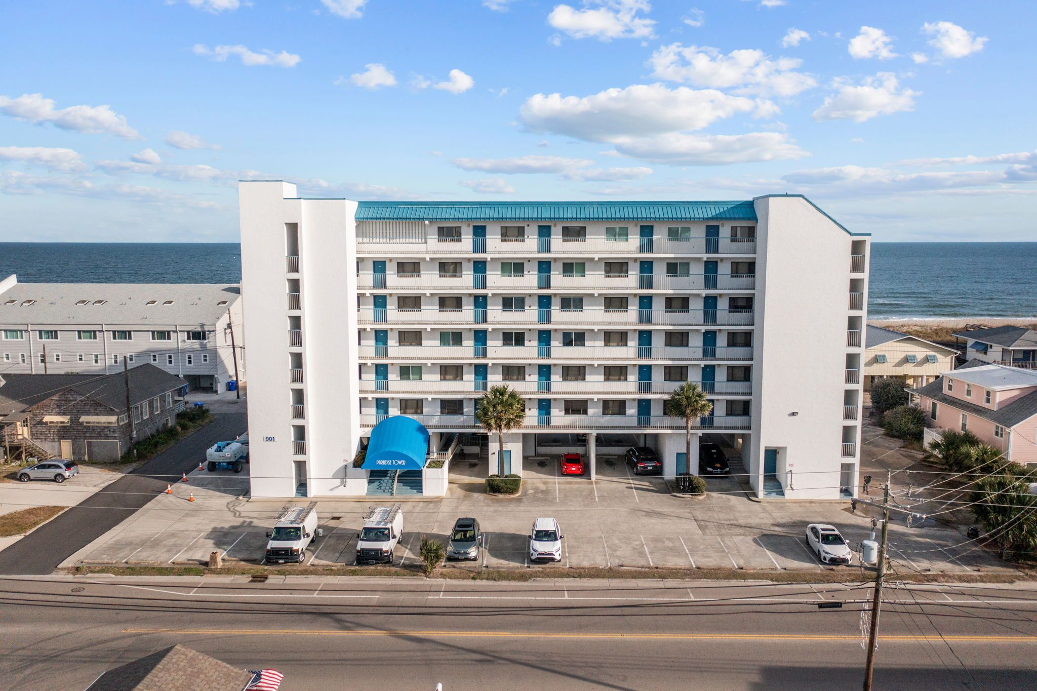 A multi-story hotel with a blue roof, parking lot, and a view of the ocean.