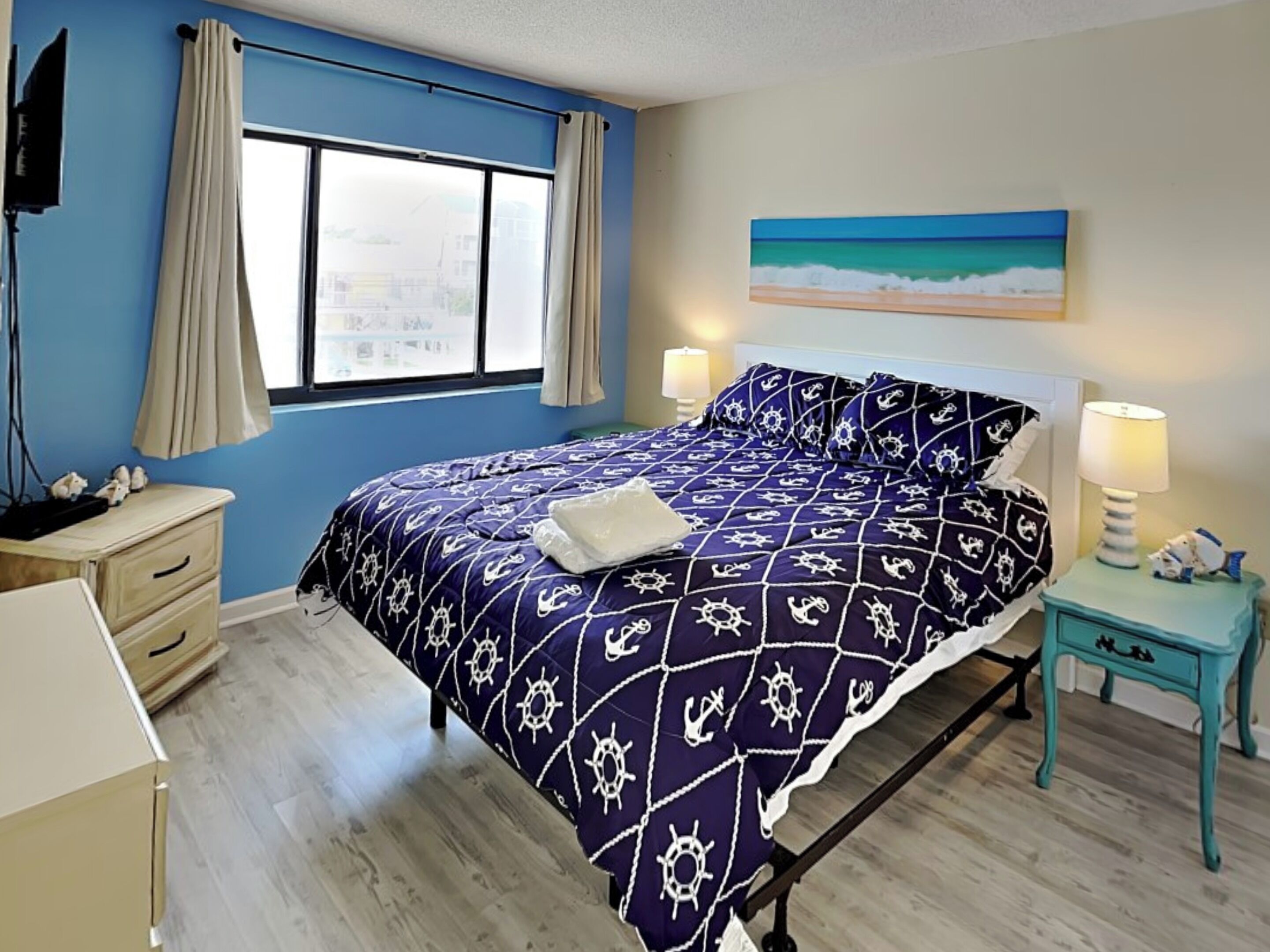 A bedroom with a bed, two bedside tables, a window with curtains, a painting on the wall, and a lamp on one of the tables.