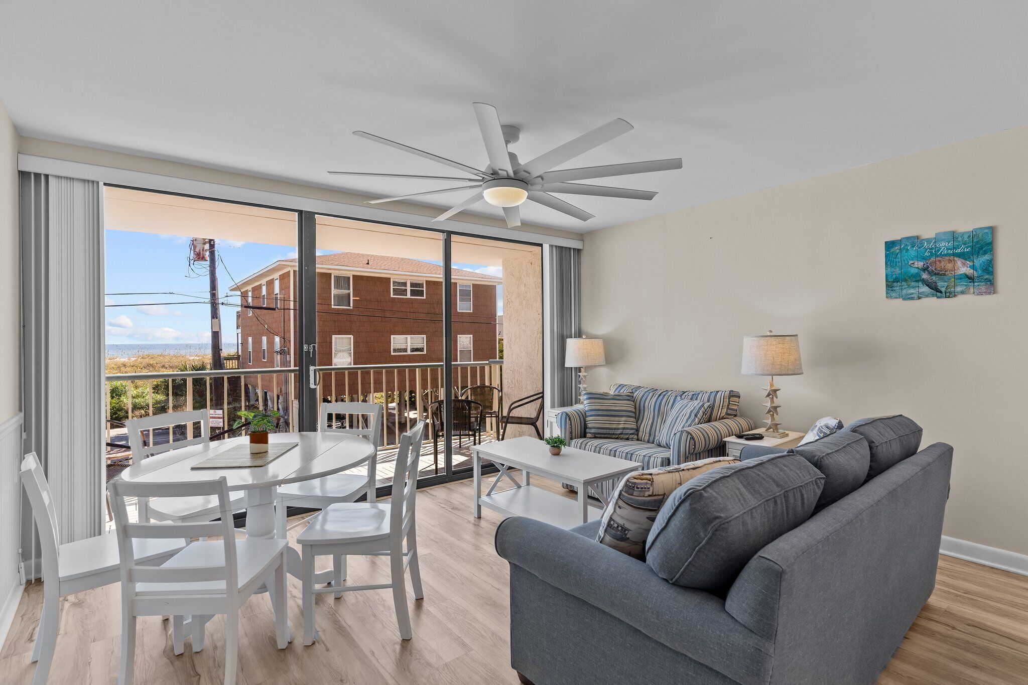 Un soggiorno moderno con un divano grigio, un tavolo da pranzo bianco e sedie. Si nota una ventola a soffitto, un'ampia finestra con vista su un edificio e un quadro appeso al muro.