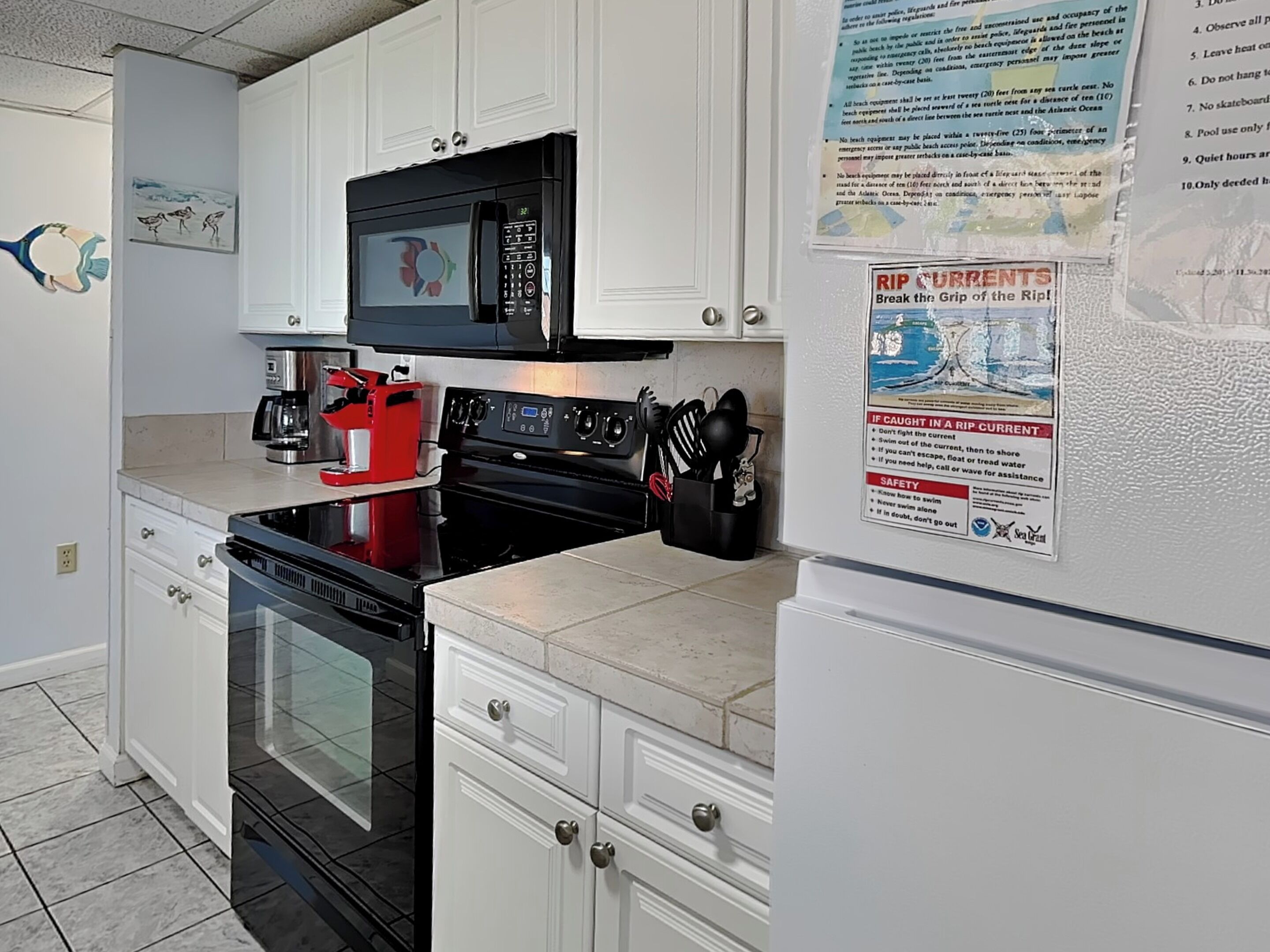 A kitchen with white cabinets, a black microwave, a red coffee maker, and a stove. There is a poster on the wall about rip currents.