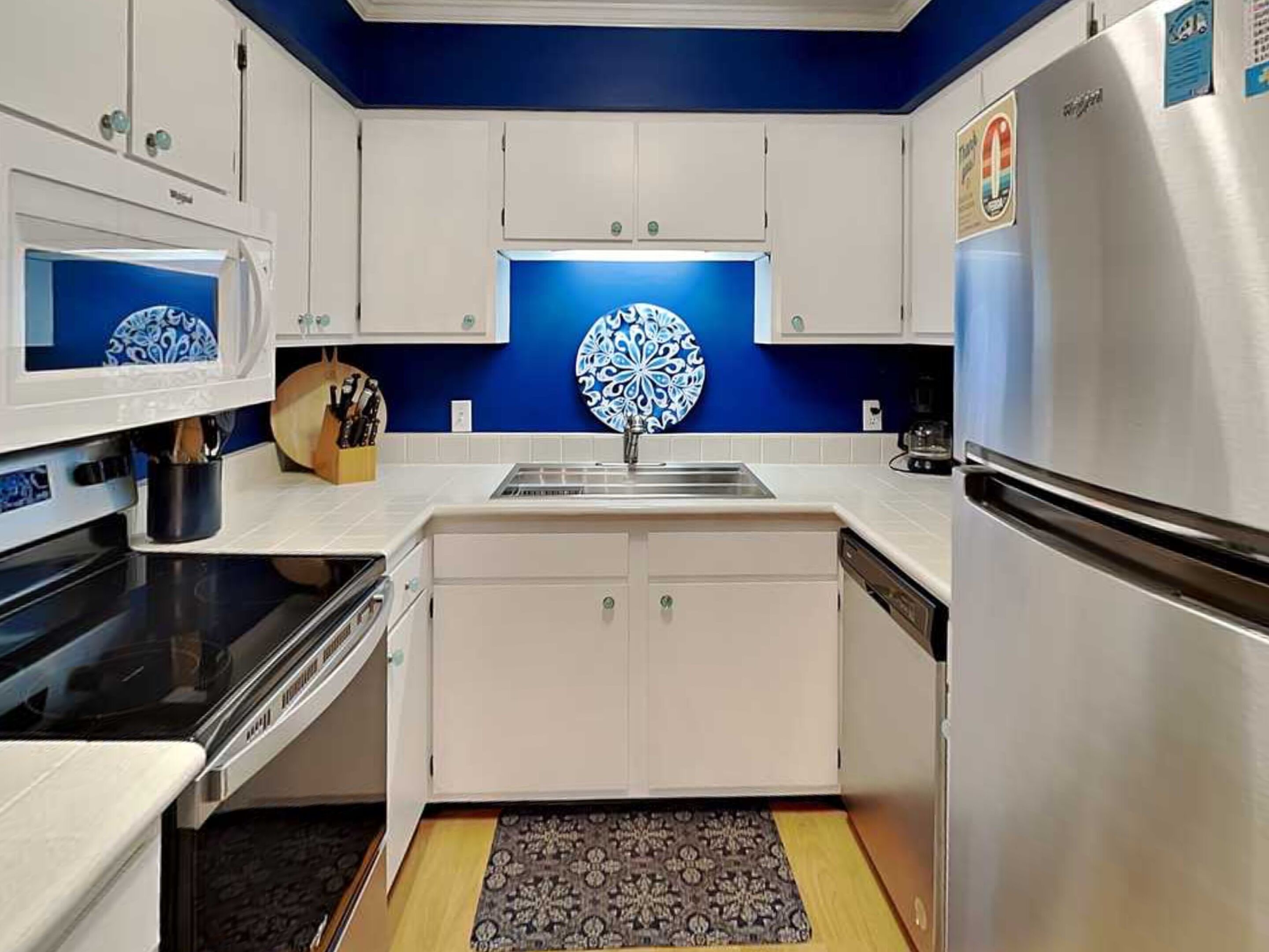 Una cucina moderna con mobili bianchi, un frigorifero in acciaio inossidabile, un forno a microonde e un lavello con pannello posteriore blu.