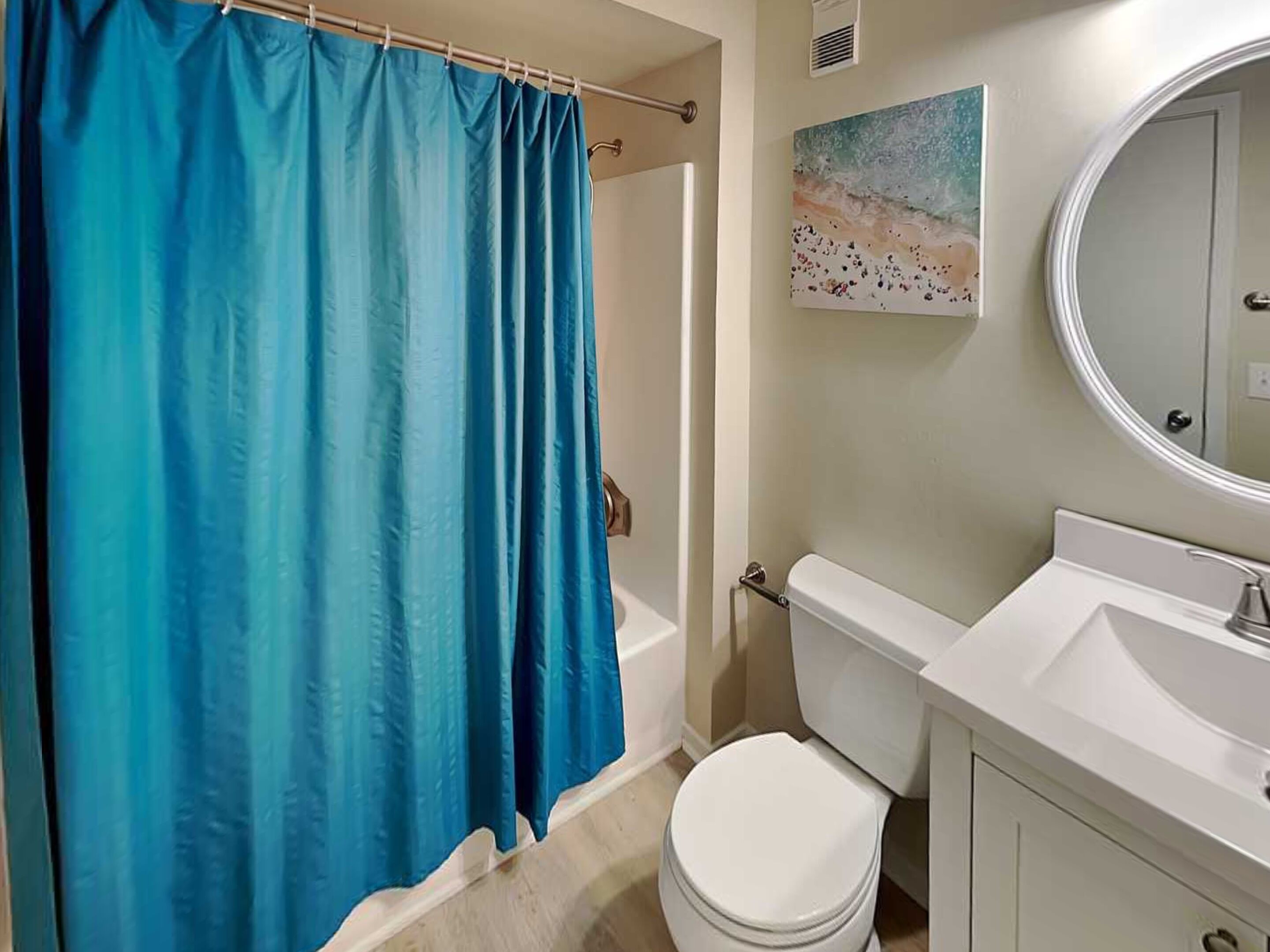A bathroom with a white toilet, a white sink, a blue shower curtain, and a painting on the wall.