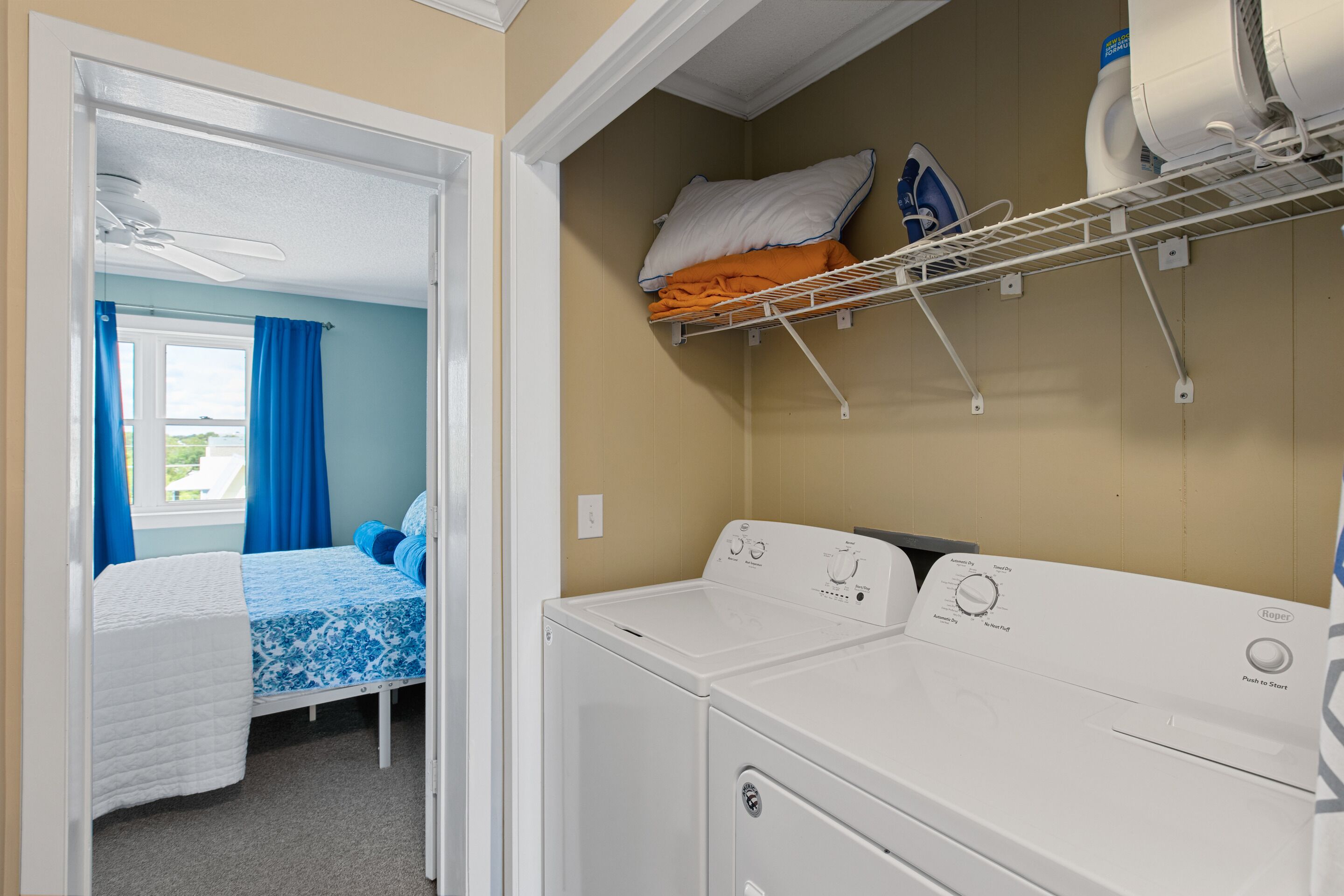 A laundry room with a washer and dryer, a bed with blue bedding, and a window with blue curtains.