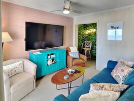 Una sala de estar con un televisor de pantalla plana grande, mueble de entretenimiento color turquesa, sillón color beige y sofá azul, junto con una pequeña mesa de centro.