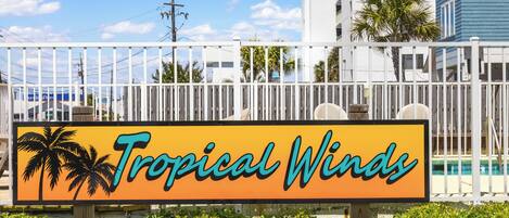 Señal de Tropical Winds con palmeras y una piscina al fondo.