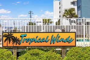 Schild für Tropical Winds, im Hintergrund Palmen und ein Pool.