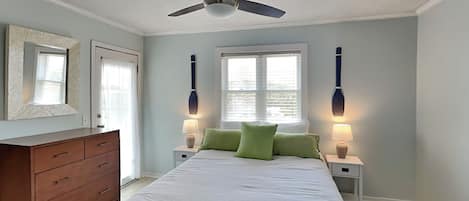 Un dormitorio con una cama, un aparador, dos mesitas de noche con lámparas, un ventilador de techo y dos ventanas con persianas.