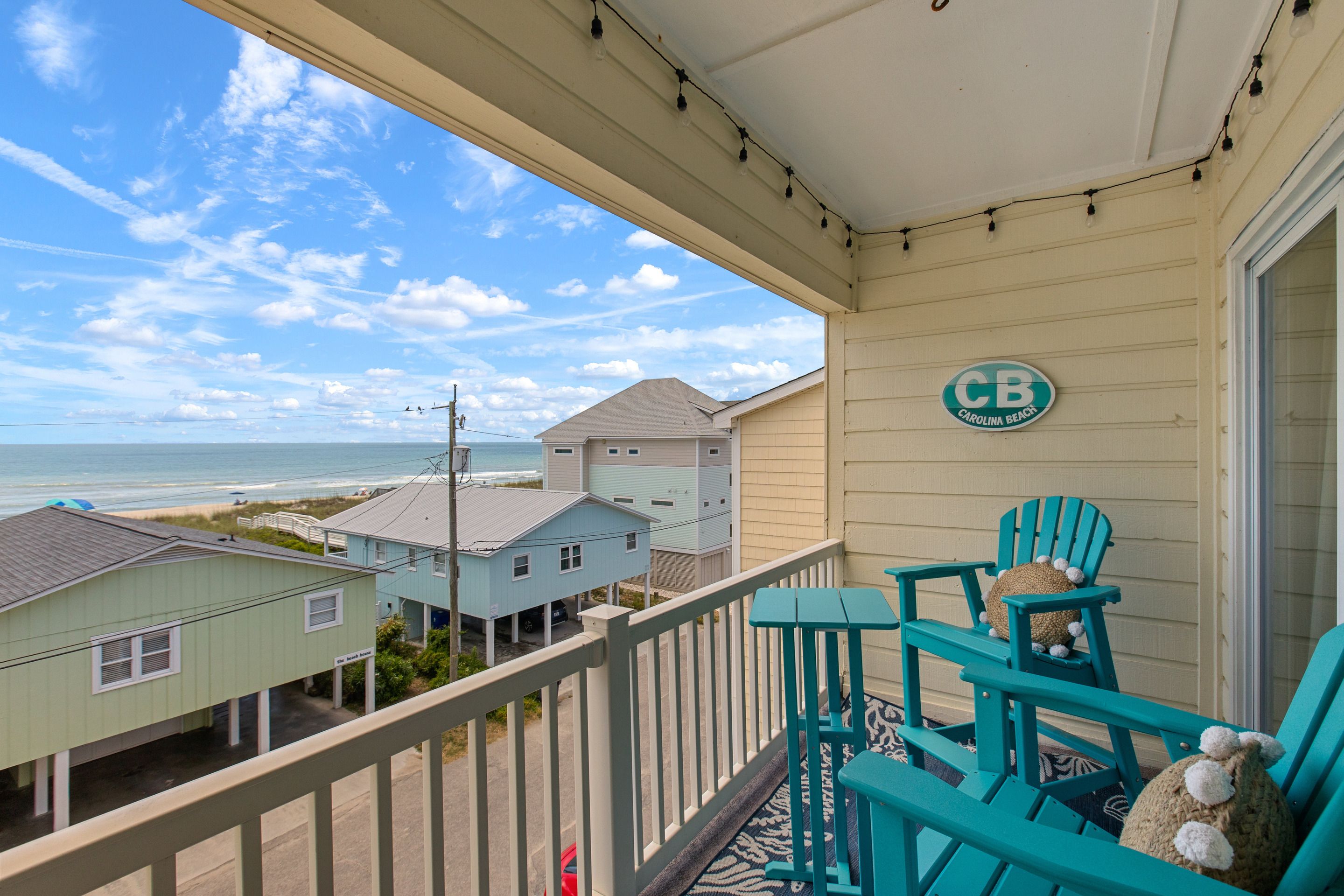 A balcony with teal chairs and a 'CB' sign, overlooking a beach and houses.