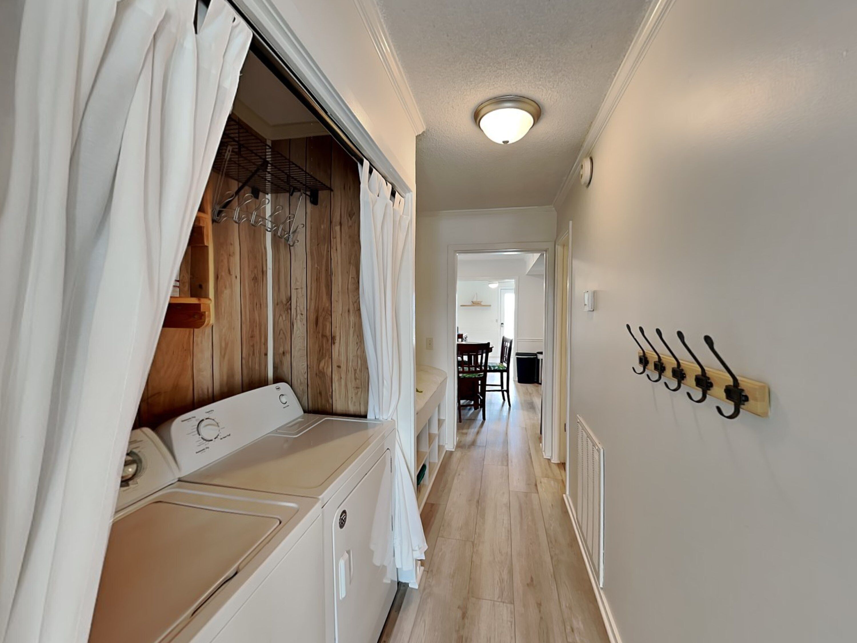 A laundry room with a washing machine, drying rack, and a coat rack. A hallway leads to a dining area.