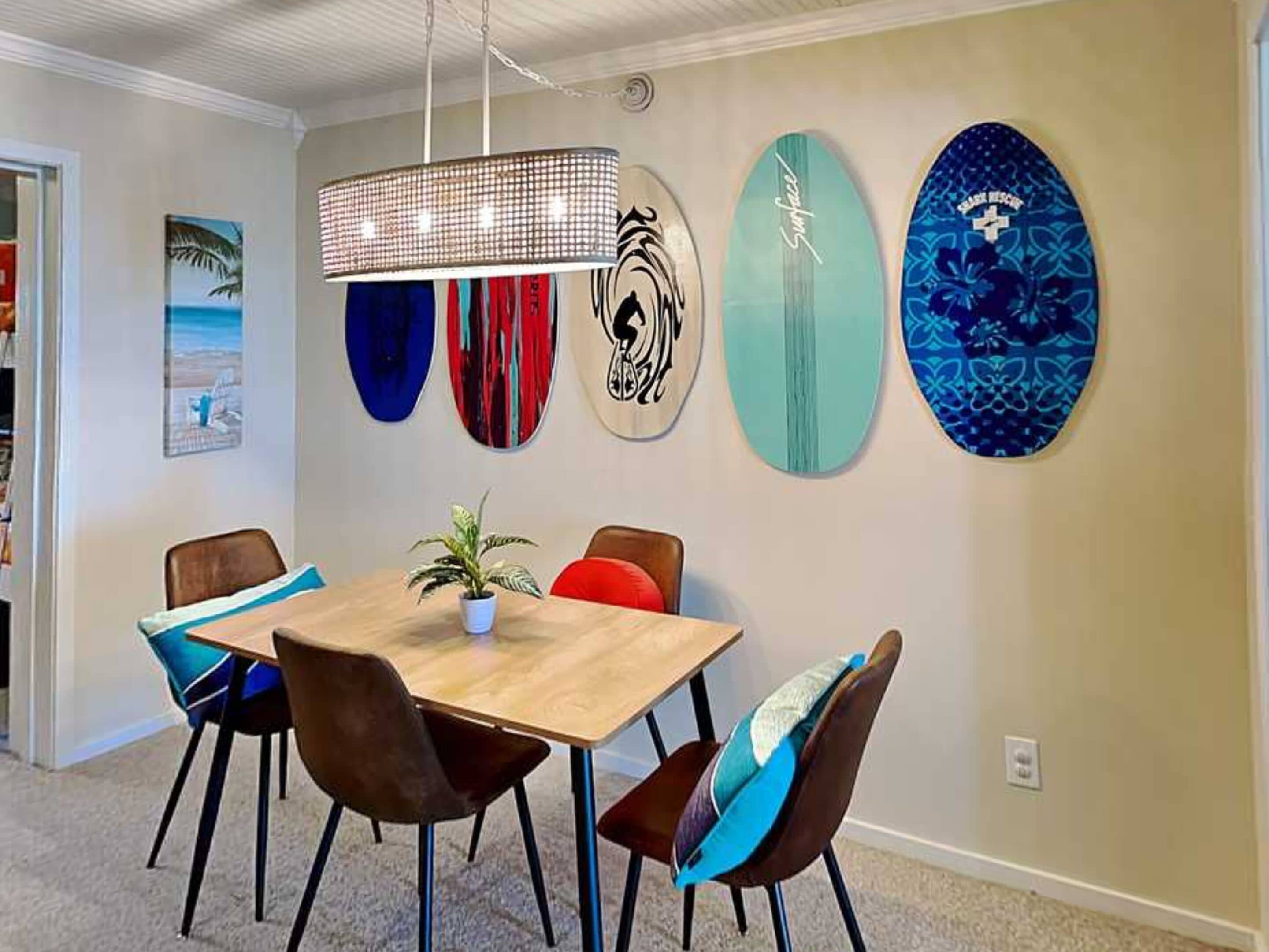 A dining area with a wooden table, four chairs, a hanging light fixture, and surfboards on the wall.