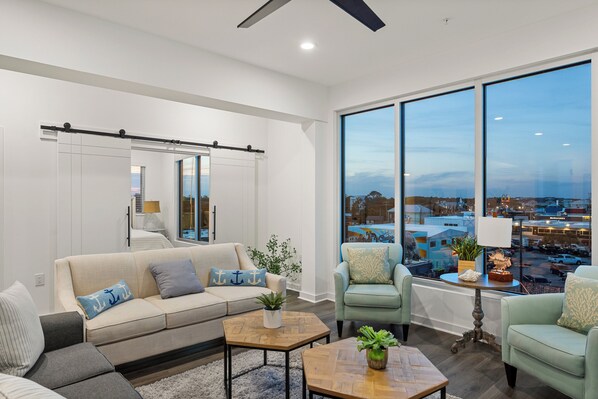 A modern living room with a large window, a sofa, two armchairs, a coffee table, and a side table with a lamp. - Boardwalk Pent House by Carolina Retreats (Carolina Beach)