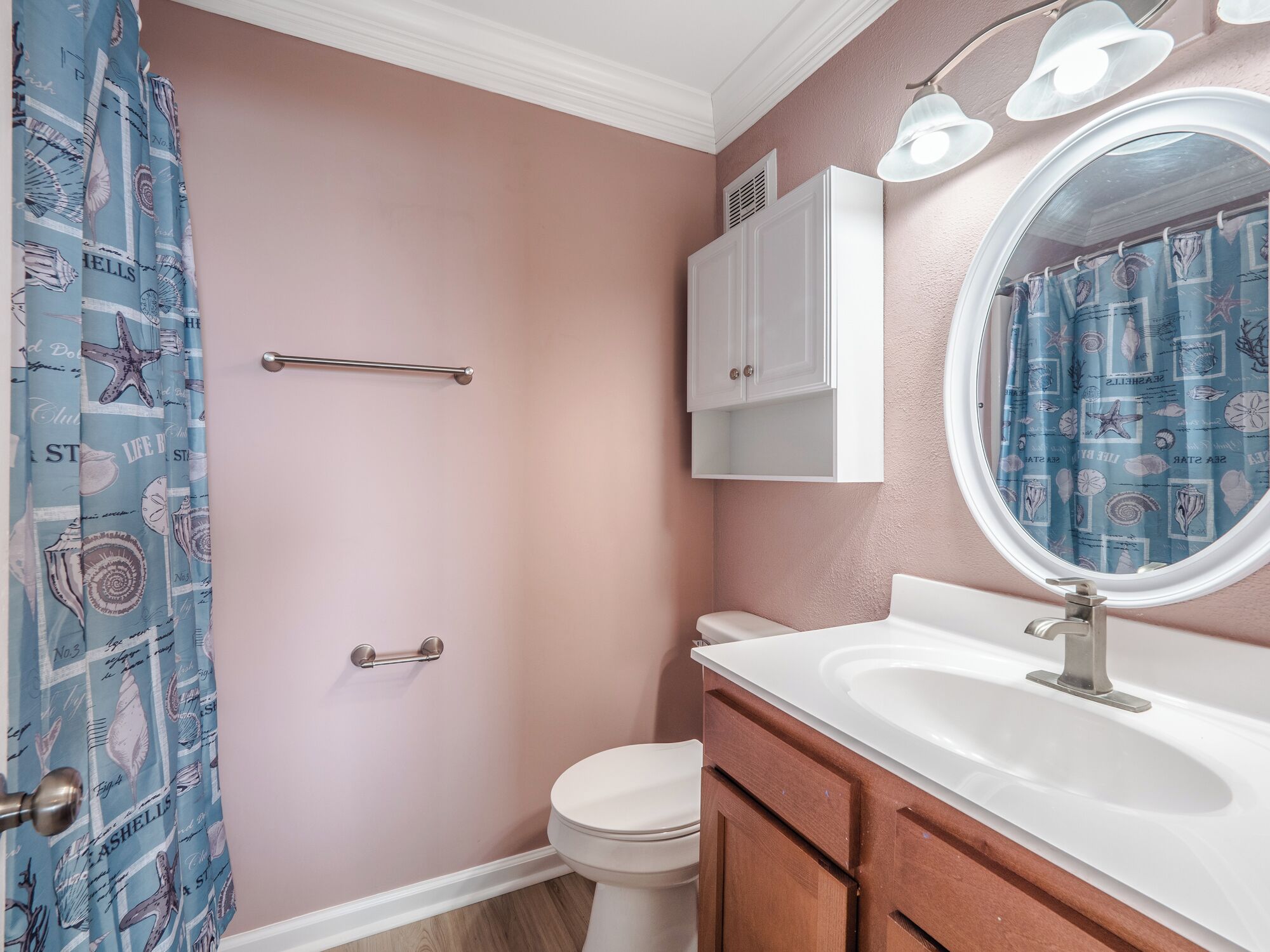 A bathroom with a pink wall, a white toilet, a wooden vanity with a sink, and a shower curtain with a nautical theme.