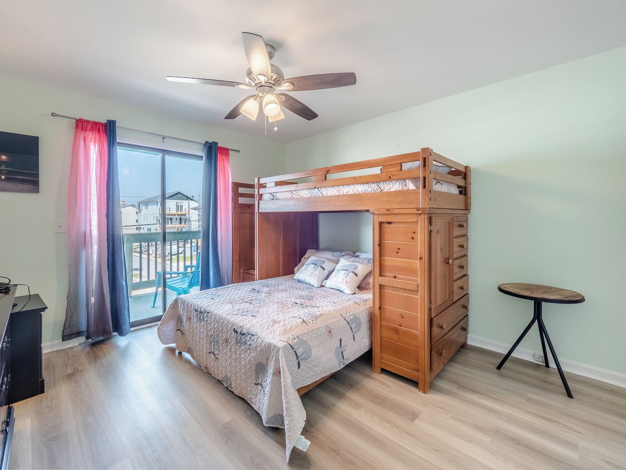 A room with a bunk bed, a ceiling fan, a small table, and a view of the outside.