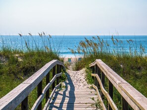 A wooden boardwalk leading to a beach with clear blue water and grassy areas. - Winds by Carolina Retreats (Carolina Beach)